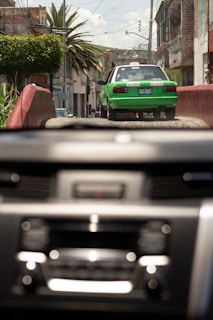 A green and white taxi drives down a rustic street with buildings and palm trees lining the sides. The image is viewed through a car windshield with a blurred dashboard in the foreground. It appears to be a bright day with a clear sky.