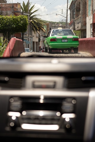 A green and white taxi drives down a rustic street with buildings and palm trees lining the sides. The image is viewed through a car windshield with a blurred dashboard in the foreground. It appears to be a bright day with a clear sky.