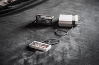A vintage gaming console setup is placed on a dark fabric surface. The setup includes a classic Nintendo Entertainment System (NES) console, a connected game controller, and a Nikon film camera nearby.