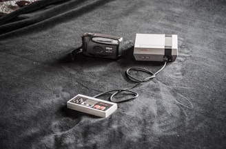 A vintage gaming console setup is placed on a dark fabric surface. The setup includes a classic Nintendo Entertainment System (NES) console, a connected game controller, and a Nikon film camera nearby.
