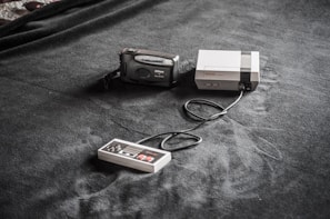 A vintage gaming console setup is placed on a dark fabric surface. The setup includes a classic Nintendo Entertainment System (NES) console, a connected game controller, and a Nikon film camera nearby.
