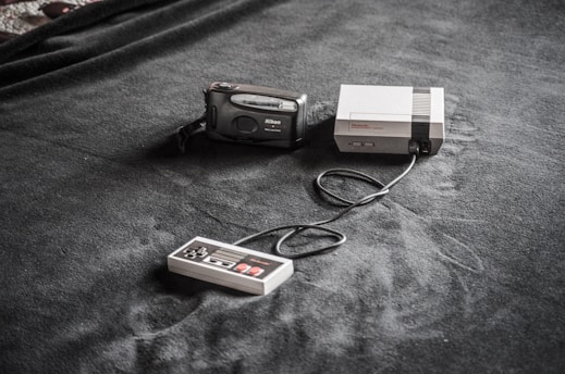 A vintage gaming console setup is placed on a dark fabric surface. The setup includes a classic Nintendo Entertainment System (NES) console, a connected game controller, and a Nikon film camera nearby.