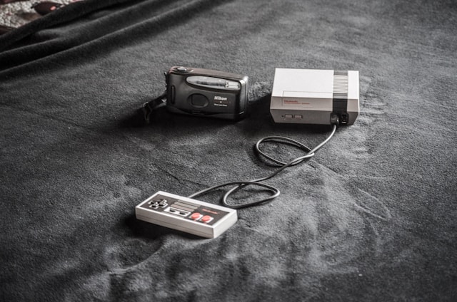 A vintage gaming console setup is placed on a dark fabric surface. The setup includes a classic Nintendo Entertainment System (NES) console, a connected game controller, and a Nikon film camera nearby.