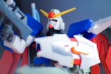 A highly detailed model of a humanoid robot with vibrant colors, featuring a white face, red chin piece, green eyes, and a prominent yellow V-shaped antenna on its head. The body is mostly blue and red, with white and gray accents, holding what appears to be a futuristic weapon in its hand. The background is a blurred dark gradient, which emphasizes the model's vivid colors.