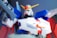 A highly detailed model of a humanoid robot with vibrant colors, featuring a white face, red chin piece, green eyes, and a prominent yellow V-shaped antenna on its head. The body is mostly blue and red, with white and gray accents, holding what appears to be a futuristic weapon in its hand. The background is a blurred dark gradient, which emphasizes the model's vivid colors.