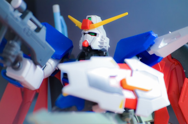 A highly detailed model of a humanoid robot with vibrant colors, featuring a white face, red chin piece, green eyes, and a prominent yellow V-shaped antenna on its head. The body is mostly blue and red, with white and gray accents, holding what appears to be a futuristic weapon in its hand. The background is a blurred dark gradient, which emphasizes the model's vivid colors.