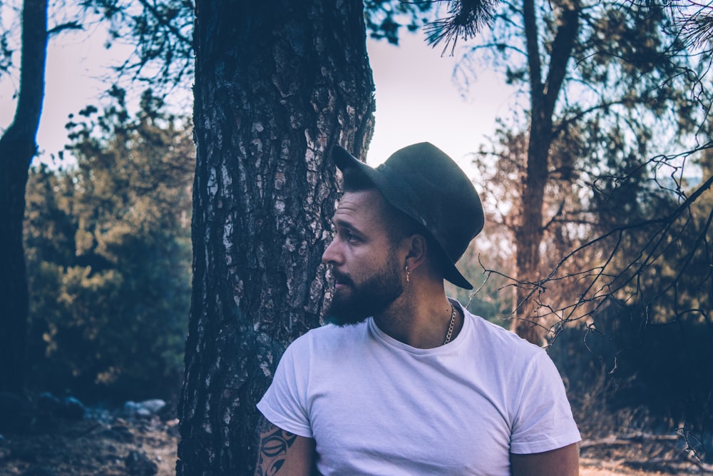 man leaning on brown tree wearing black fedora hat and white crew-neck shirt looking to side