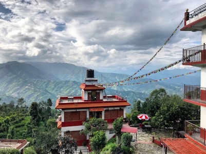 A serene mountain landscape with a multi-story building featuring red accents and a rooftop water tank. Colorful prayer flags are strung between the buildings, adding a cultural touch. Verdant hills and distant mountains are shrouded in mist, under a sky filled with dramatic clouds. A red and white striped umbrella shades a small seating area on the stone-paved ground.