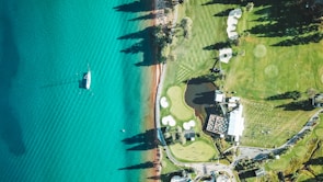 An aerial view of a golf course next to a large, blue body of water. The scene includes well-maintained greens, sand traps, and a clubhouse. A sailboat is visible on the water, casting a shadow. The landscape features trees and clear skies, adding depth to the scenery.