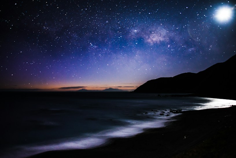 A serene night sky filled with stars stretches over a calm ocean shoreline with silhouetted hills. The bright moon illuminates the scene, casting reflections on the water. The horizon shows a subtle gradient of twilight colors, transitioning from deep blues to soft oranges.
