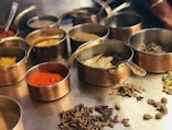 A variety of spices are displayed in several copper bowls with handles, arranged on a metal surface. The spices include vibrant red chili powder, turmeric, cardamom pods, bay leaves, and other dried herbs. The scene suggests a culinary preparation setting with an emphasis on aromatic ingredients.