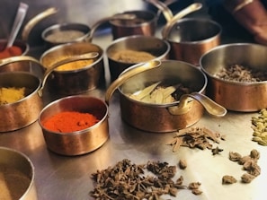 A variety of spices are displayed in several copper bowls with handles, arranged on a metal surface. The spices include vibrant red chili powder, turmeric, cardamom pods, bay leaves, and other dried herbs. The scene suggests a culinary preparation setting with an emphasis on aromatic ingredients.
