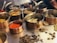 A variety of spices are displayed in several copper bowls with handles, arranged on a metal surface. The spices include vibrant red chili powder, turmeric, cardamom pods, bay leaves, and other dried herbs. The scene suggests a culinary preparation setting with an emphasis on aromatic ingredients.