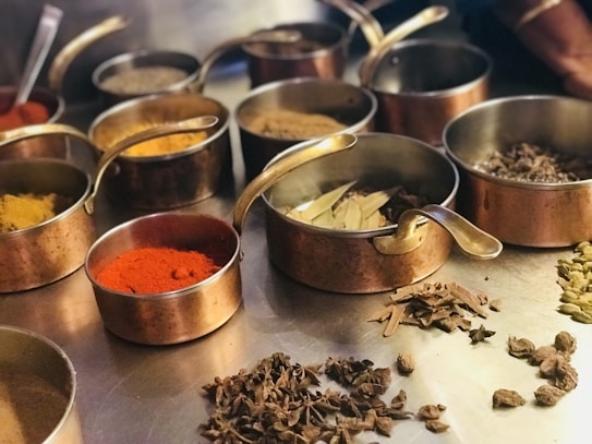 A variety of spices are displayed in several copper bowls with handles, arranged on a metal surface. The spices include vibrant red chili powder, turmeric, cardamom pods, bay leaves, and other dried herbs. The scene suggests a culinary preparation setting with an emphasis on aromatic ingredients.