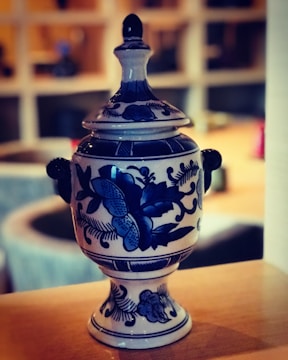 A decorative ceramic urn with intricate blue floral patterns and a lid, positioned on a wooden surface. The background is softly blurred, suggesting an indoor setting with warm lighting.