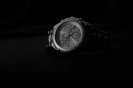 A metallic wristwatch with a multi-dial face is placed against a dark background. The watch has a detailed and classic design, featuring several subdials and a silver metal link strap.