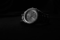 A metallic wristwatch with a multi-dial face is placed against a dark background. The watch has a detailed and classic design, featuring several subdials and a silver metal link strap.