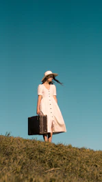 woman standing on green grass holding brown leather suitcase wearing white button-up dress