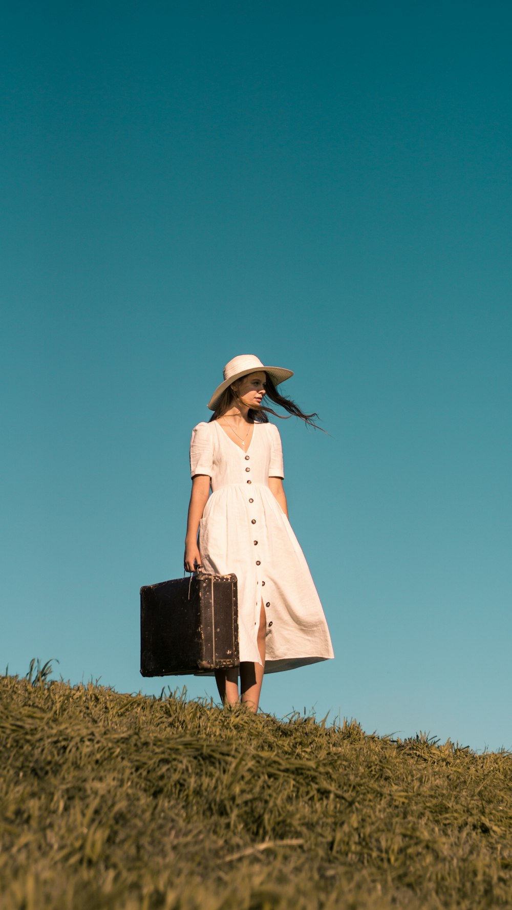 白いボタンアップのドレスを着た茶色の革のスーツケースを持って緑の芝生の上に立つ女性