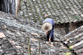A person wearing a wide-brimmed straw hat is working on repairing or inspecting a roof made up of traditional grey tiles. The roof tiles are arranged in an overlapping pattern and show various shades of grey, with patches of moss or discoloration.
