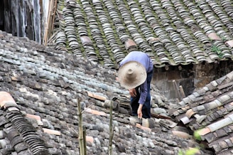 A person wearing a wide-brimmed straw hat is working on repairing or inspecting a roof made up of traditional grey tiles. The roof tiles are arranged in an overlapping pattern and show various shades of grey, with patches of moss or discoloration.