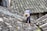 A person wearing a wide-brimmed straw hat is working on repairing or inspecting a roof made up of traditional grey tiles. The roof tiles are arranged in an overlapping pattern and show various shades of grey, with patches of moss or discoloration.