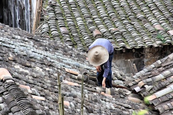A person wearing a wide-brimmed straw hat is working on repairing or inspecting a roof made up of traditional grey tiles. The roof tiles are arranged in an overlapping pattern and show various shades of grey, with patches of moss or discoloration.
