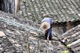 A person wearing a wide-brimmed straw hat is working on repairing or inspecting a roof made up of traditional grey tiles. The roof tiles are arranged in an overlapping pattern and show various shades of grey, with patches of moss or discoloration.