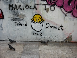 A piece of street art on a wall features a cartoonish drawing of a yellow chick partially emerged from a half eggshell. Above the chick is the word 'Friend' and below it is 'not Omelet'. The wall is decorated with various graffiti and the ground shows three pigeons walking.