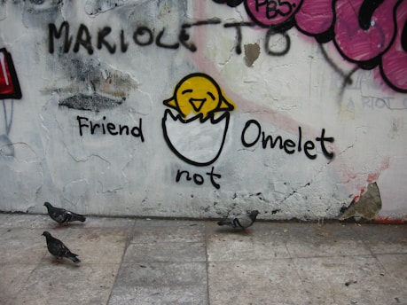 A piece of street art on a wall features a cartoonish drawing of a yellow chick partially emerged from a half eggshell. Above the chick is the word 'Friend' and below it is 'not Omelet'. The wall is decorated with various graffiti and the ground shows three pigeons walking.