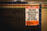 A newspaper poster is attached to a wall with colorful tiles and an old plaque. The headline reads 'West End Final: Trump Launches Missile Strike At Syria,' with the publication being the London Evening Standard.