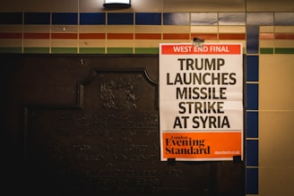 A newspaper poster is attached to a wall with colorful tiles and an old plaque. The headline reads 'West End Final: Trump Launches Missile Strike At Syria,' with the publication being the London Evening Standard.