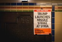 A newspaper poster is attached to a wall with colorful tiles and an old plaque. The headline reads 'West End Final: Trump Launches Missile Strike At Syria,' with the publication being the London Evening Standard.
