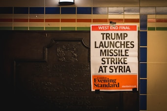 A newspaper poster is attached to a wall with colorful tiles and an old plaque. The headline reads 'West End Final: Trump Launches Missile Strike At Syria,' with the publication being the London Evening Standard.