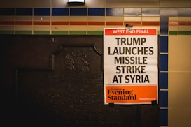 A newspaper poster is attached to a wall with colorful tiles and an old plaque. The headline reads 'West End Final: Trump Launches Missile Strike At Syria,' with the publication being the London Evening Standard.