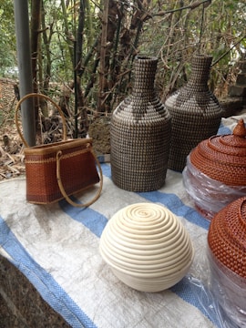 Several woven objects are displayed on a surface with a blue and white checkered cloth. There are two tall, intricately patterned bottles, a small brown woven handbag with a round handle, and a spherical basket with a coiled white design in the foreground. Additionally, two conical-shaped lids or baskets are partly wrapped in plastic. The background features dense, lush bamboo foliage.