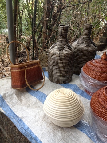 Several woven objects are displayed on a surface with a blue and white checkered cloth. There are two tall, intricately patterned bottles, a small brown woven handbag with a round handle, and a spherical basket with a coiled white design in the foreground. Additionally, two conical-shaped lids or baskets are partly wrapped in plastic. The background features dense, lush bamboo foliage.