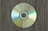 A compact disc lies on a textured surface, displaying a spectrum of colors reflected across its shiny surface. The background consists of a fabric-like pattern with earth-toned colors, providing a contrast to the vibrant hues on the disc.