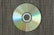A compact disc lies on a textured surface, displaying a spectrum of colors reflected across its shiny surface. The background consists of a fabric-like pattern with earth-toned colors, providing a contrast to the vibrant hues on the disc.