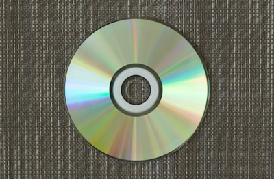 A compact disc lies on a textured surface, displaying a spectrum of colors reflected across its shiny surface. The background consists of a fabric-like pattern with earth-toned colors, providing a contrast to the vibrant hues on the disc.