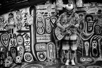 A person stands against a vibrantly decorated wall covered in abstract and tribal-style paintings and symbols. The individual wears a patterned shirt, knee pads, and a helmet resembling the Iron Man mask. They are playing a guitar adorned with similar ornate designs found on the wall. The scene is engaging and filled with artistic expressions.