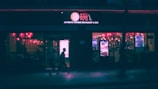 A dimly lit Chinese restaurant and bar is visible at night. Red lanterns hang inside, casting a warm glow through the windows. The street outside is relatively dark, with silhouettes of people walking by.