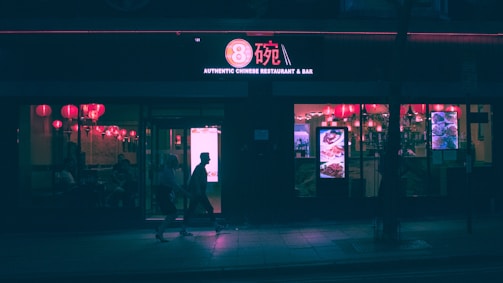 A dimly lit Chinese restaurant and bar is visible at night. Red lanterns hang inside, casting a warm glow through the windows. The street outside is relatively dark, with silhouettes of people walking by.