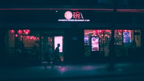 A dimly lit Chinese restaurant and bar is visible at night. Red lanterns hang inside, casting a warm glow through the windows. The street outside is relatively dark, with silhouettes of people walking by.