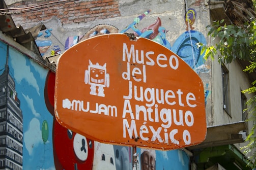 An orange sign with white text that reads 'Museo del Juguete Antiguo México' featuring a cartoon robot graphic. The sign is surrounded by colorful murals on adjacent walls, depicting various vibrant art pieces. There is a mix of urban and artistic elements with visible greenery.