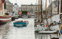A lively harbor scene with numerous boats docked along a canal. In the foreground, a blue tour boat filled with people is cruising down the water. On either side of the canal, there are old brick buildings and vibrant storefronts, including visible flags. The atmosphere suggests a bustling and popular tourist area.