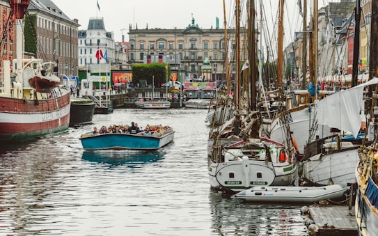A lively harbor scene with numerous boats docked along a canal. In the foreground, a blue tour boat filled with people is cruising down the water. On either side of the canal, there are old brick buildings and vibrant storefronts, including visible flags. The atmosphere suggests a bustling and popular tourist area.