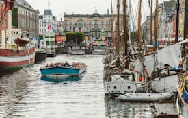 A lively harbor scene with numerous boats docked along a canal. In the foreground, a blue tour boat filled with people is cruising down the water. On either side of the canal, there are old brick buildings and vibrant storefronts, including visible flags. The atmosphere suggests a bustling and popular tourist area.