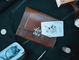 A leather-bound journal with an intricate patterned cover is placed atop a dark fabric surface. A playing card with a joker and flower design is partially tucked into the journal. Surrounding the journal are various objects including a silver coin, an eyeglass case with glasses inside, a pen, a wine cork, a wristwatch, and an orange box with text partially visible.