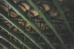 A series of green-painted steel beams intersect, creating a geometric pattern against a backdrop of brick. The structure appears robust and industrial, suggesting the underside of a bridge or a large building framework. The brickwork adds a rustic and textured element to the scene.